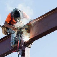 An Hispanic steel worker working high up on a girder. He is sitting on the girder, wearing a safety harness, welding to secure the girder to a column.