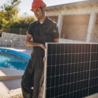 Two people, male engineers with protective helmets carrying solar panel. Professional electricians working on installation of solar panels,