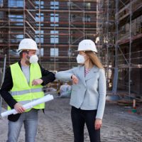 An architect or civil engineer greets his customer with an alternative greeting during the corona pandemic on the large construction site and both wear hard hats and FFP-2 masks. An apartment building can be seen in the background. Photographed in high resolution with copy space